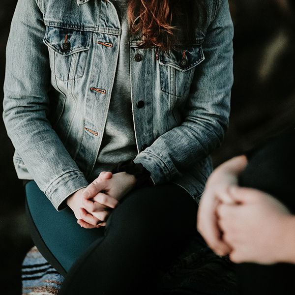 female visible from shoulders down sitting with clasped hands in lap with another female figure in foreground out of focus - unsplash image by priscilla du preez